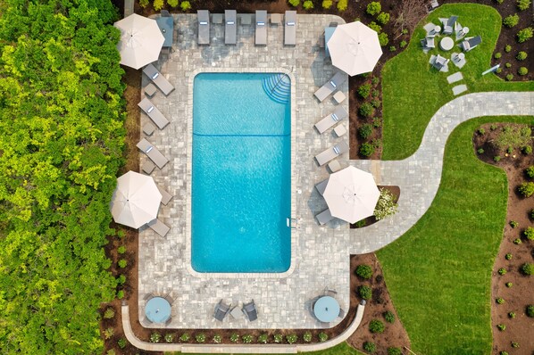 Une vue aérienne d’une piscine rectangulaire entourée de chaises longues et de parasols, avec un chemin pavé et des espaces aménagés.