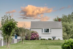 Une maison blanche avec un toit gris, entourée d’une pelouse bien entretenue et d’arbustes en fleurs.