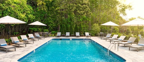 Uma piscina retangular com esguicho revestido, cercada por espreguiçadeiras e guarda-sóis, com árvores verdes densas ao fundo.