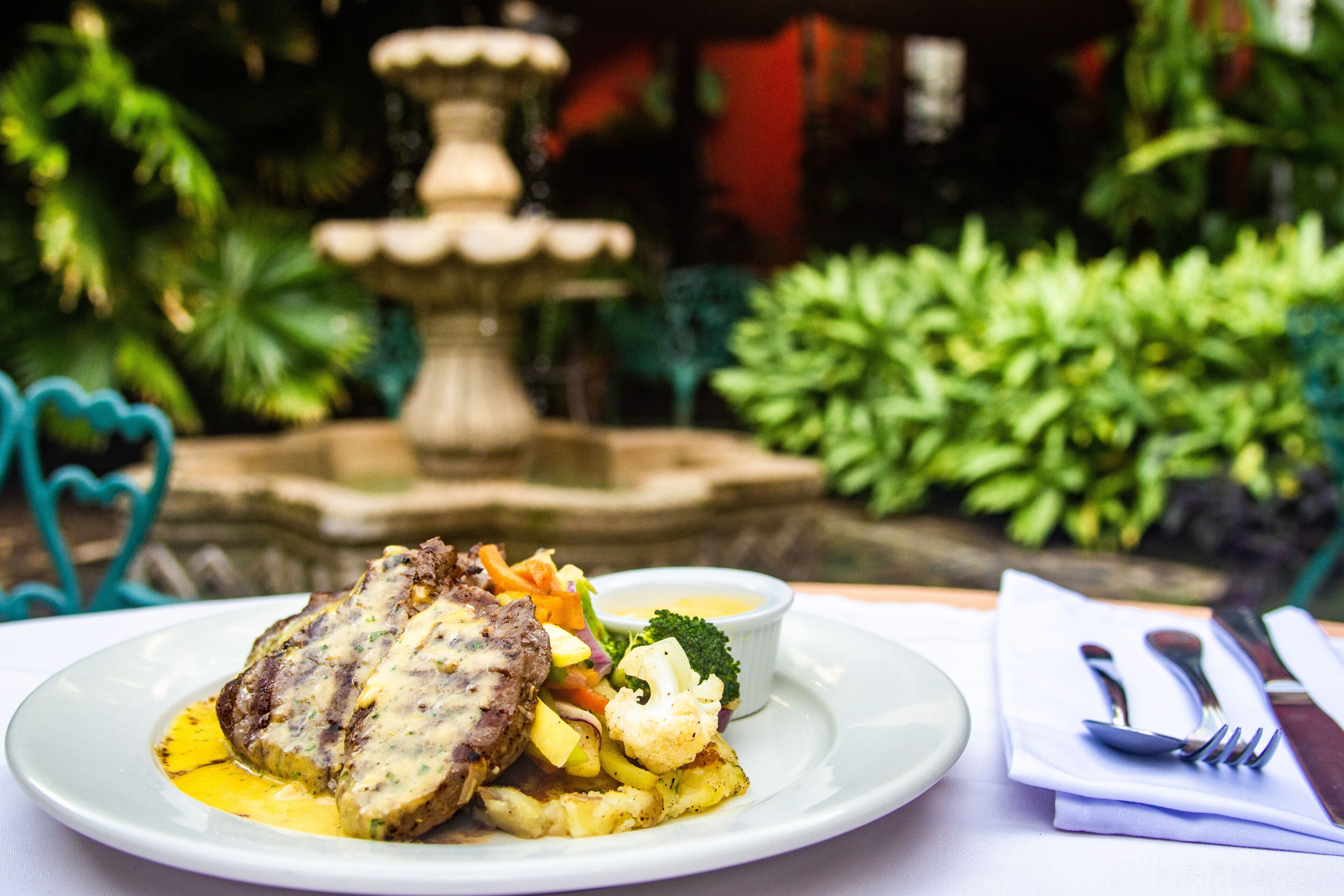 A plate of food with a meat dish, vegetables, and a small bowl of sauce, set on a table with a napkin and cutlery.