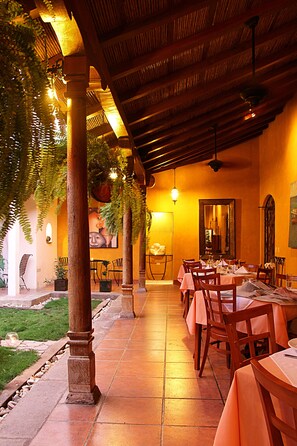 Un área de comedor al aire libre con mesas y sillas, piso de baldosas y una estructura de pérgola.