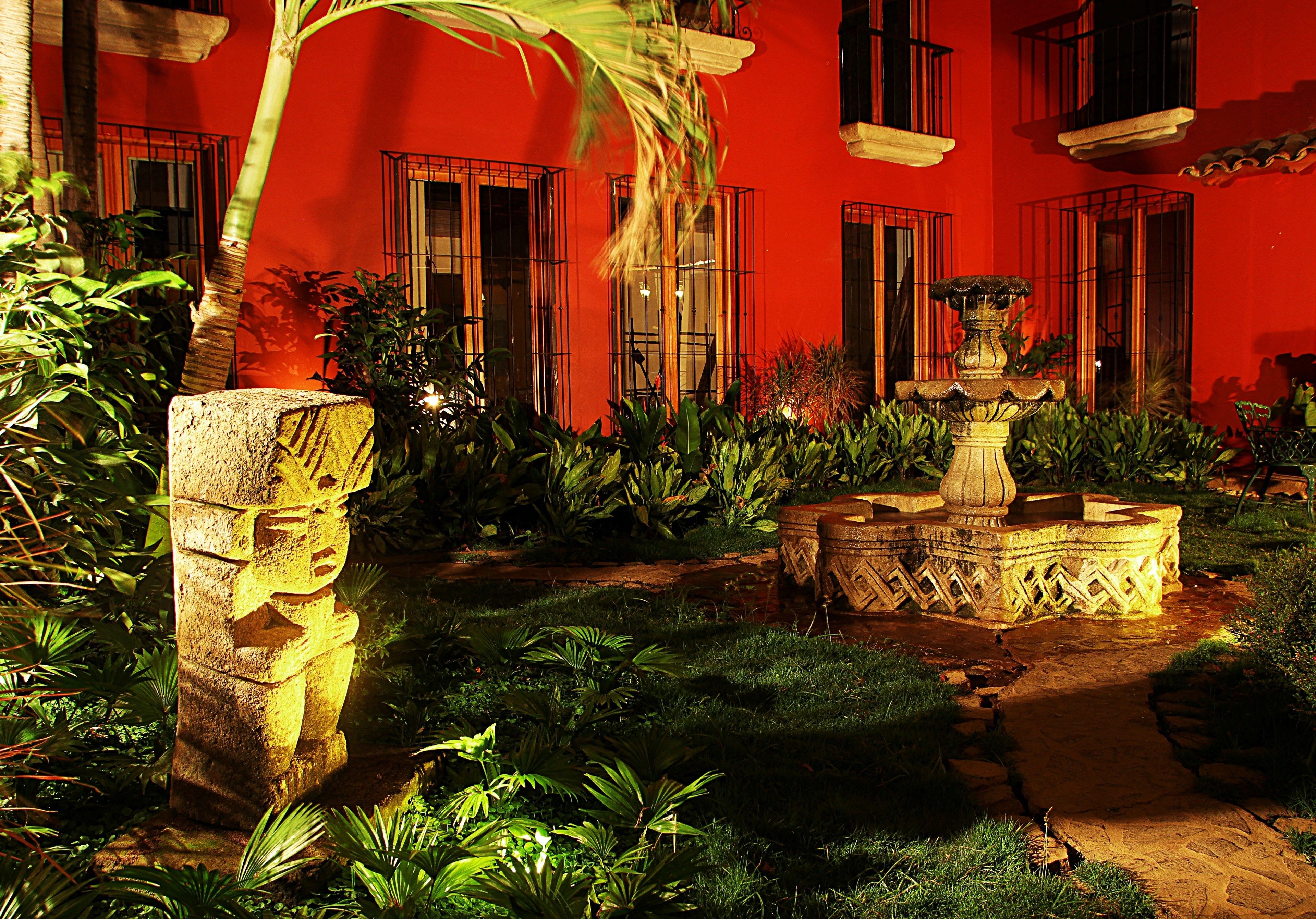 Un patio con una estatua de piedra, una fuente y abundante vegetación.