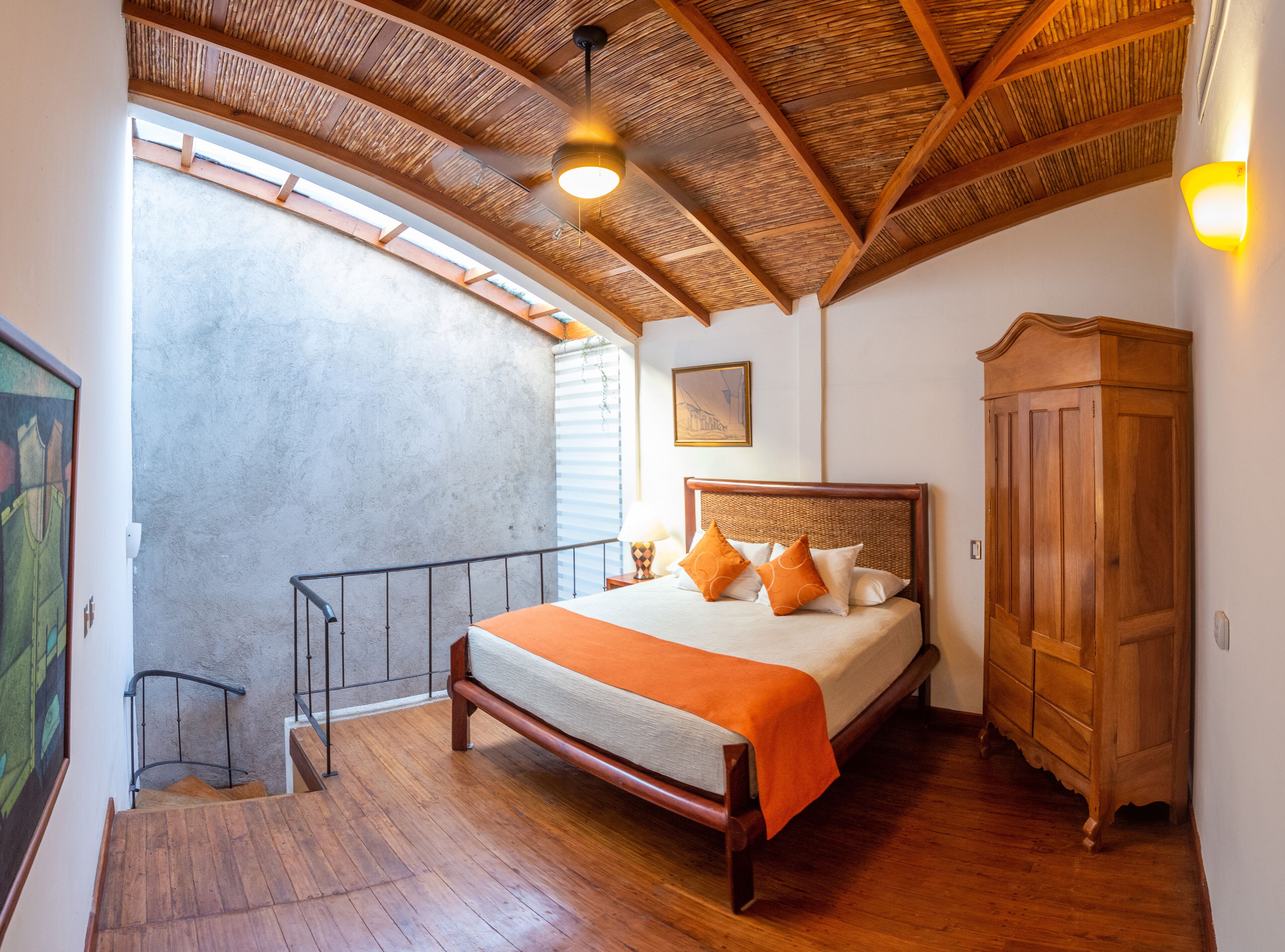 A bedroom with a wooden bed, a wardrobe, a balcony with a railing, and a ceiling with wooden beams.