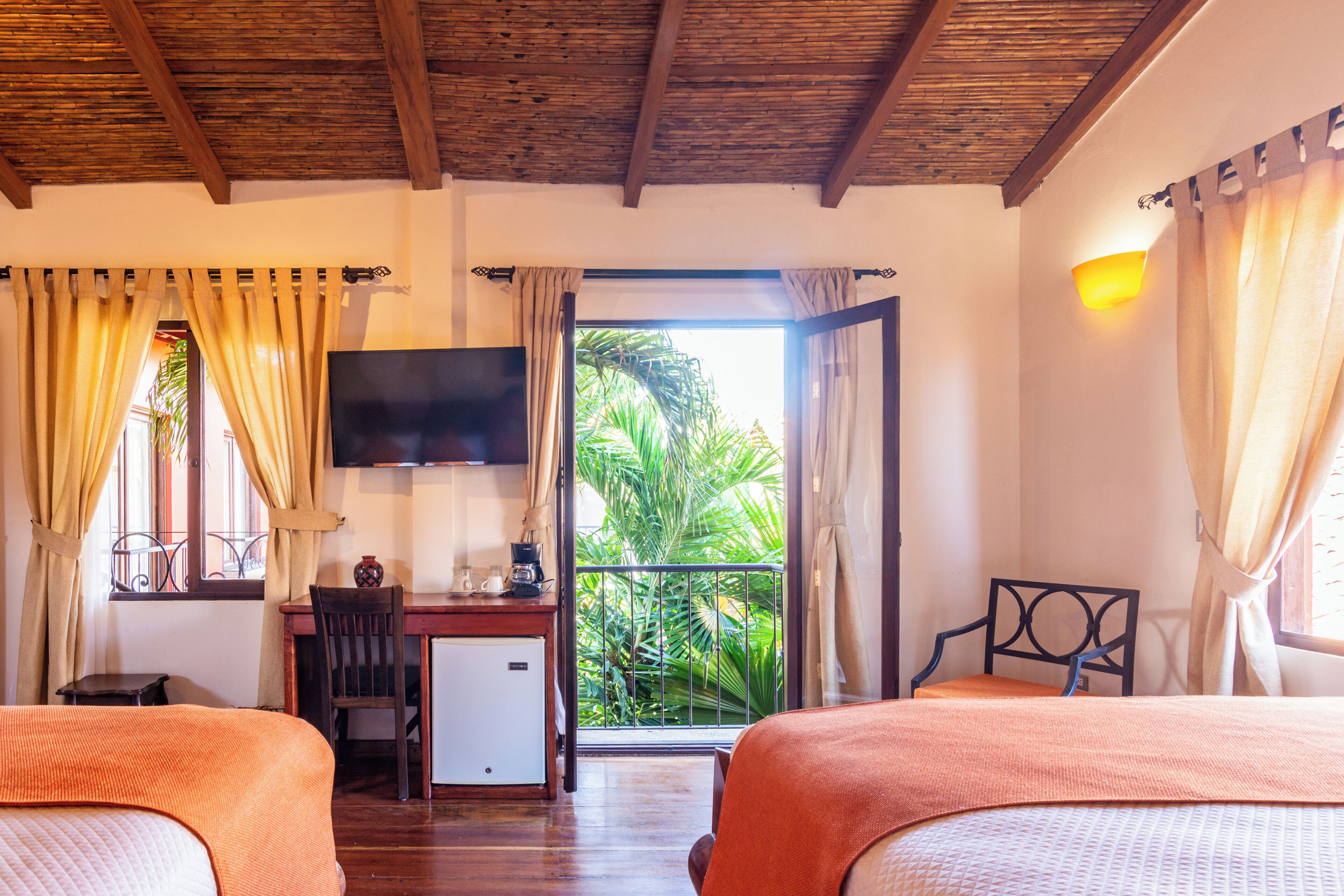 A hotel room with two beds, a desk, a TV, and a balcony with a view of greenery.