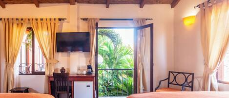 A hotel room with two beds, a desk, a TV, and a balcony with a view of greenery.