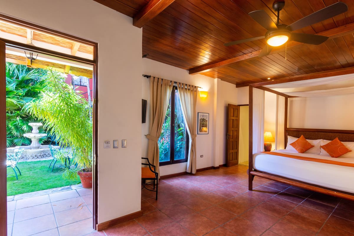 A hotel room with a bed, a ceiling fan, a window with curtains, and a view of a garden with a fountain.
