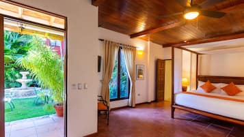 Habitación de hotel con cama, ventilador de techo, ventana con cortinas y vista a un jardín con fuente.
