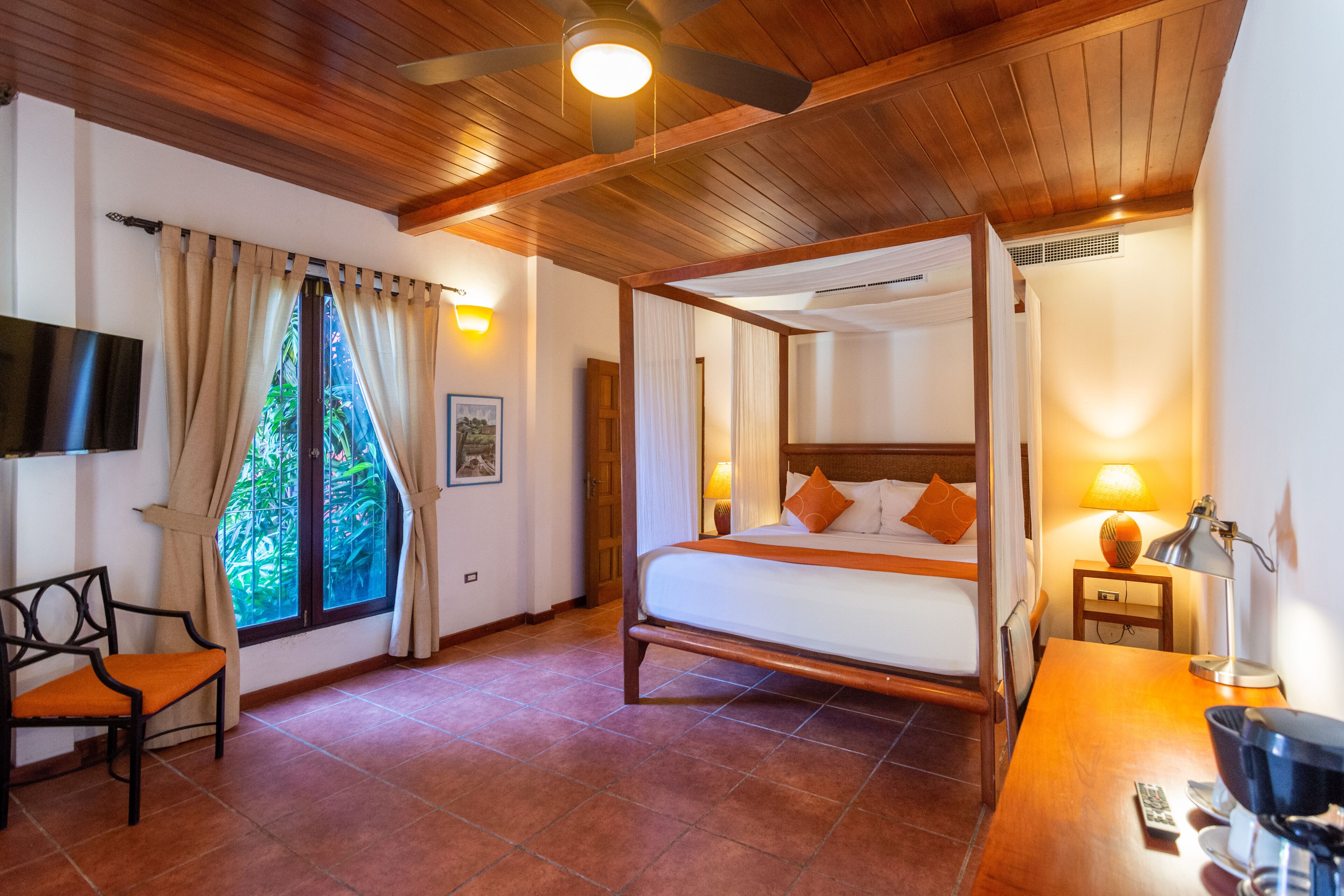 A four-poster bed with a wooden frame, a desk with a lamp, a chair, and a television on the wall.