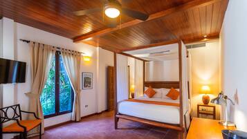 A four-poster bed with a wooden frame, a desk with a lamp, a chair, and a television on the wall.
