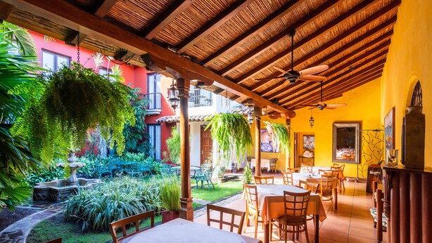 An outdoor patio with wooden tables and chairs, surrounded by lush greenery and a thatched roof. - Estrada Hotel & Bistro (Granada)