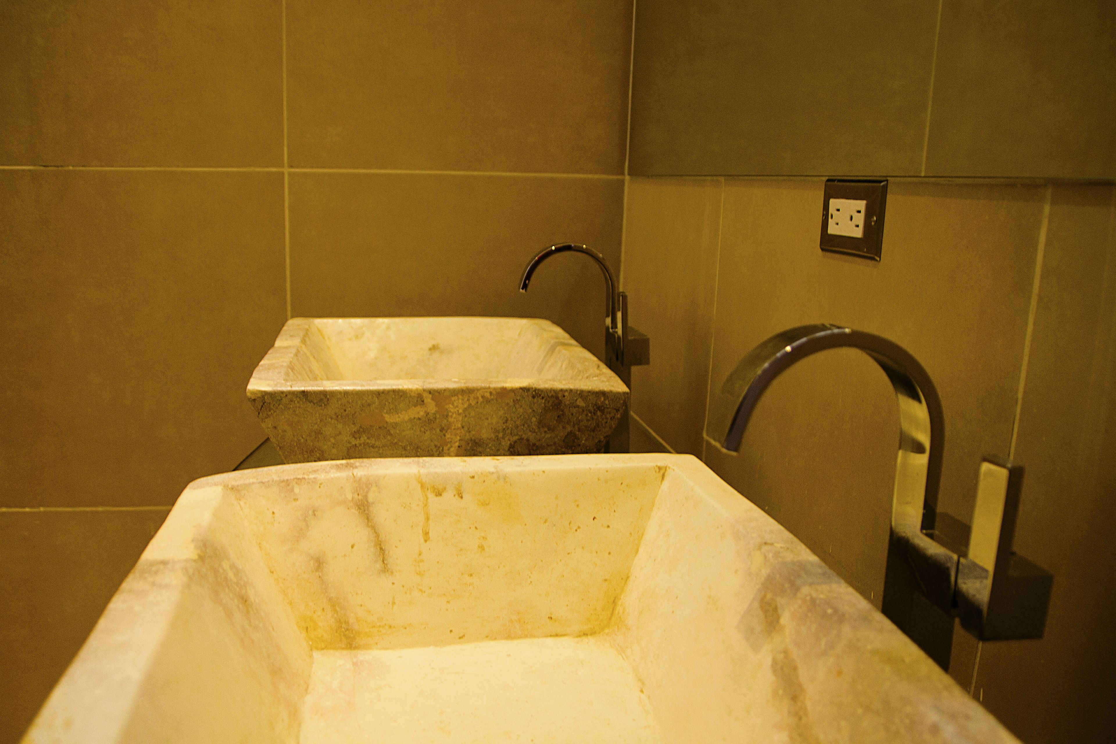 A bathroom with two sinks, one rectangular and one square, both made of a light-colored stone. There is a single faucet on the right side, and a power outlet is visible on the wall above the sinks.