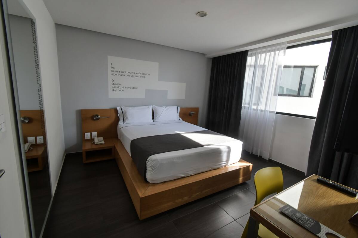 Ett modernt hotellrum med en säng i trä, ett sängbord, en stol och ett skrivbord med en fjärrkontroll.