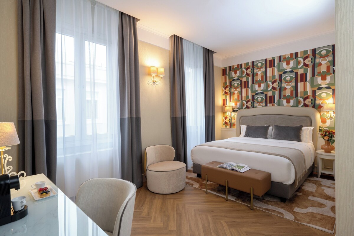 A hotel room with a large bed, a round chair, a small table with a lamp, and a wall with abstract art.
