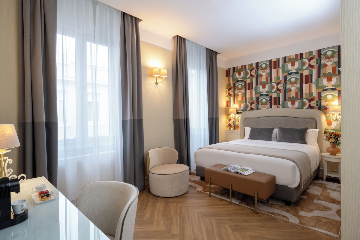 A hotel room with a large bed, a round chair, a small table with a cup and a plate of fruit, and a wall with a geometric pattern.