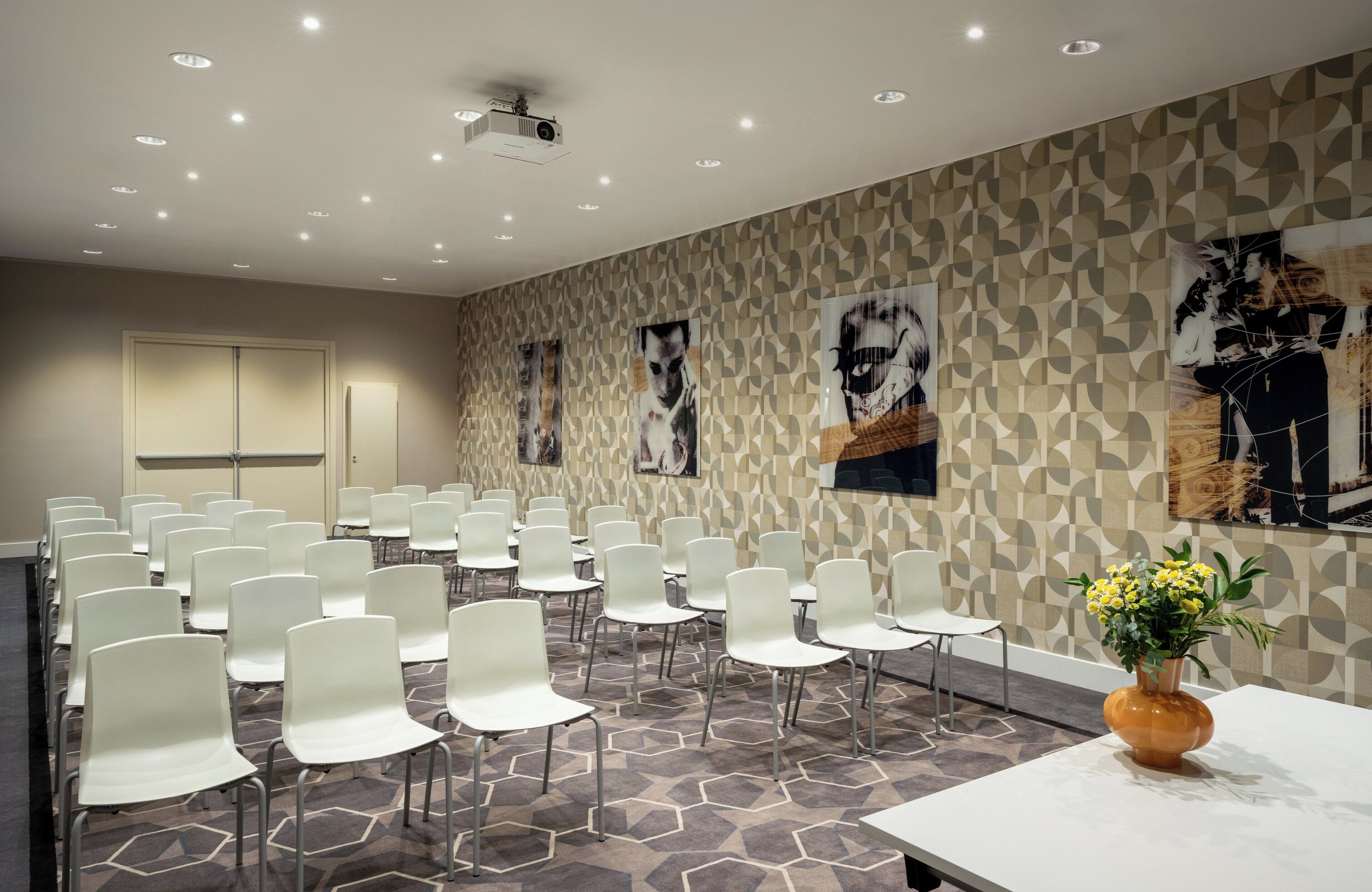 A conference room with white chairs arranged in rows, a projector on the ceiling, and a patterned wall with artwork.