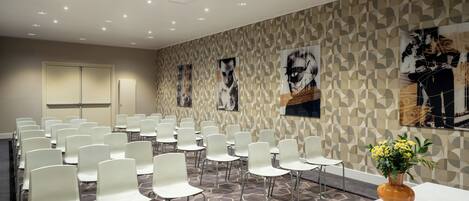 A conference room with white chairs arranged in rows, a projector on the ceiling, and a patterned wall with artwork.