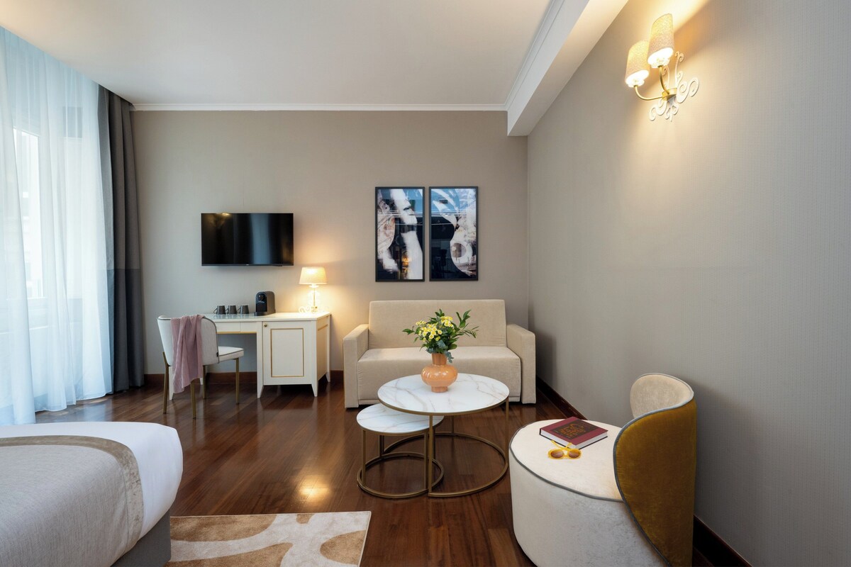 A modern hotel room with a bed, a desk, a sofa, a round table, and artwork on the walls.