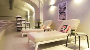 Een wellnessruimte met twee lounge fauteuils, een klein tafeltje, een plank met handdoeken, een vaas met bloemen en een schilderij aan de muur.