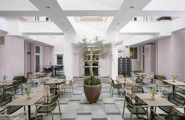 A spacious indoor dining area with multiple tables and chairs, a central large planter with a tree, and a high ceiling with skylights.