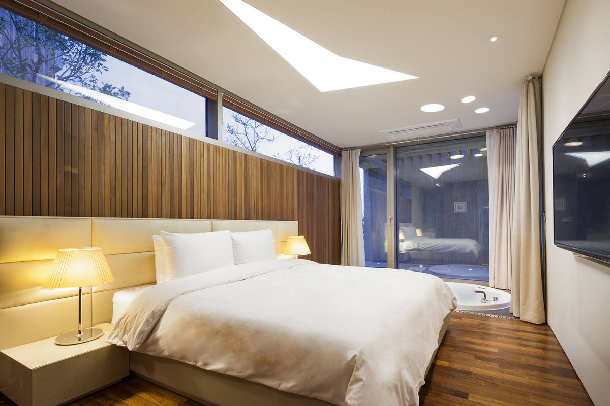 A modern hotel room with a large bed, bedside lamps, a view of the outdoors through a sliding door, and a skylight.