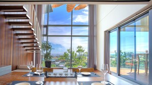 A modern dining area with a wooden table set for four, large windows offering a view of the outdoors, and a staircase with wooden steps. - Bayhill Pool & Villa (Seogwipo)