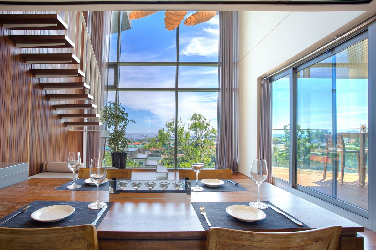 A modern dining area with a wooden table set for four, large windows offering a view of the outdoors, and a staircase with wooden steps.