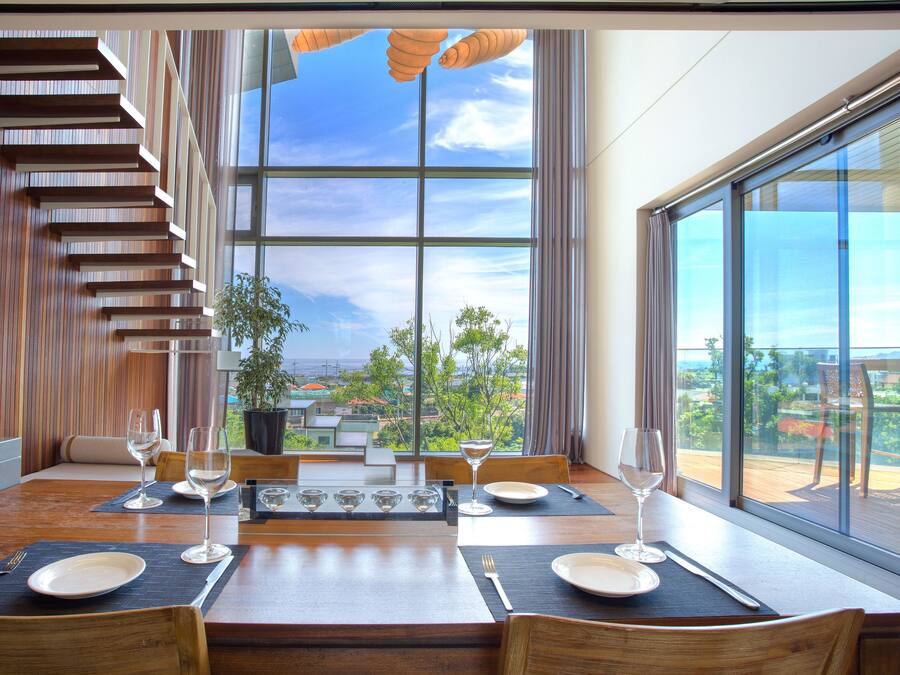 Un espace repas moderne avec une table en bois disposée pour quatre personnes, de grandes fenêtres offrant une vue sur l’extérieur et un escalier avec des marches en bois.