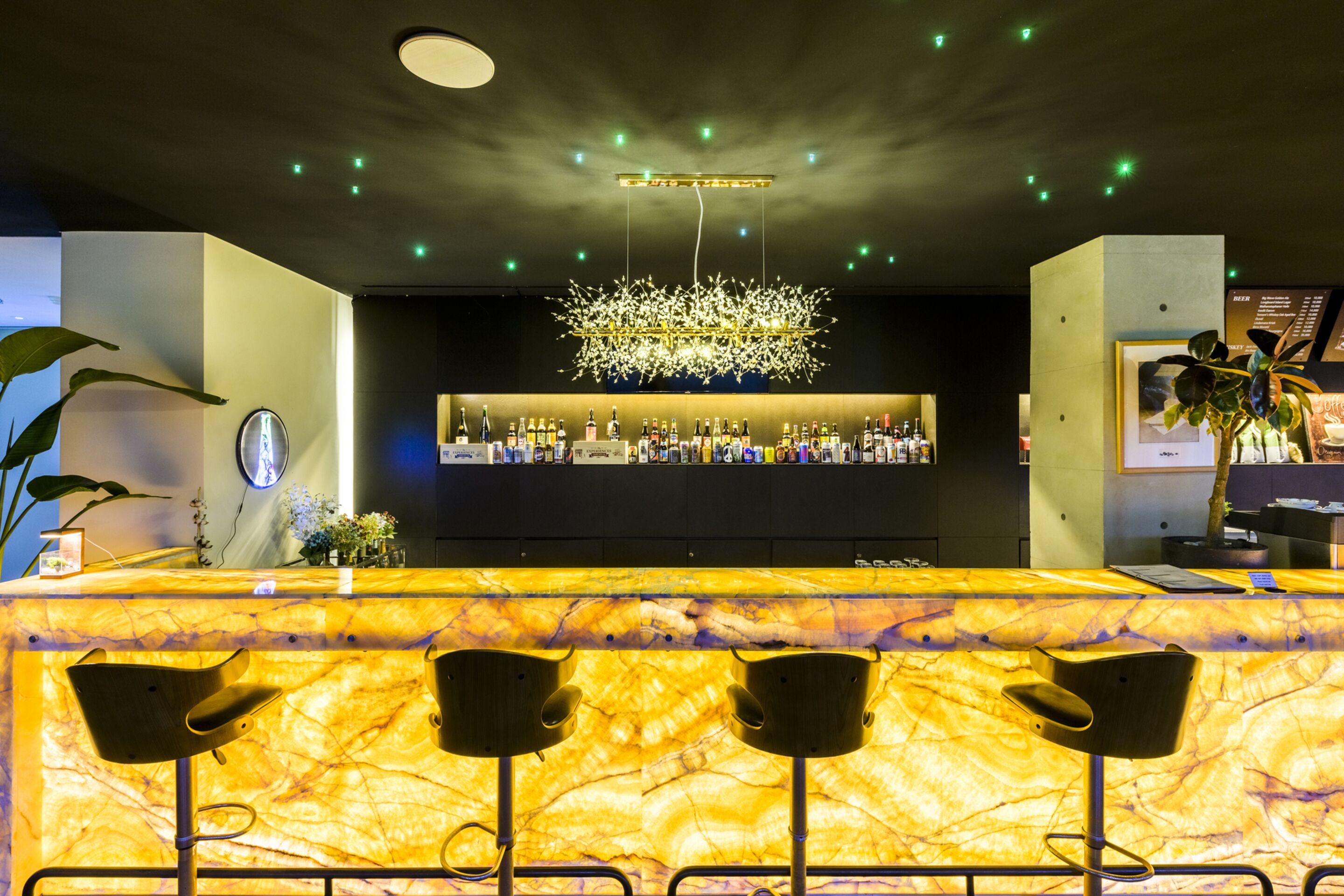 A bar area with a marble countertop, three bar stools, a chandelier, and a well-stocked shelf with bottles.