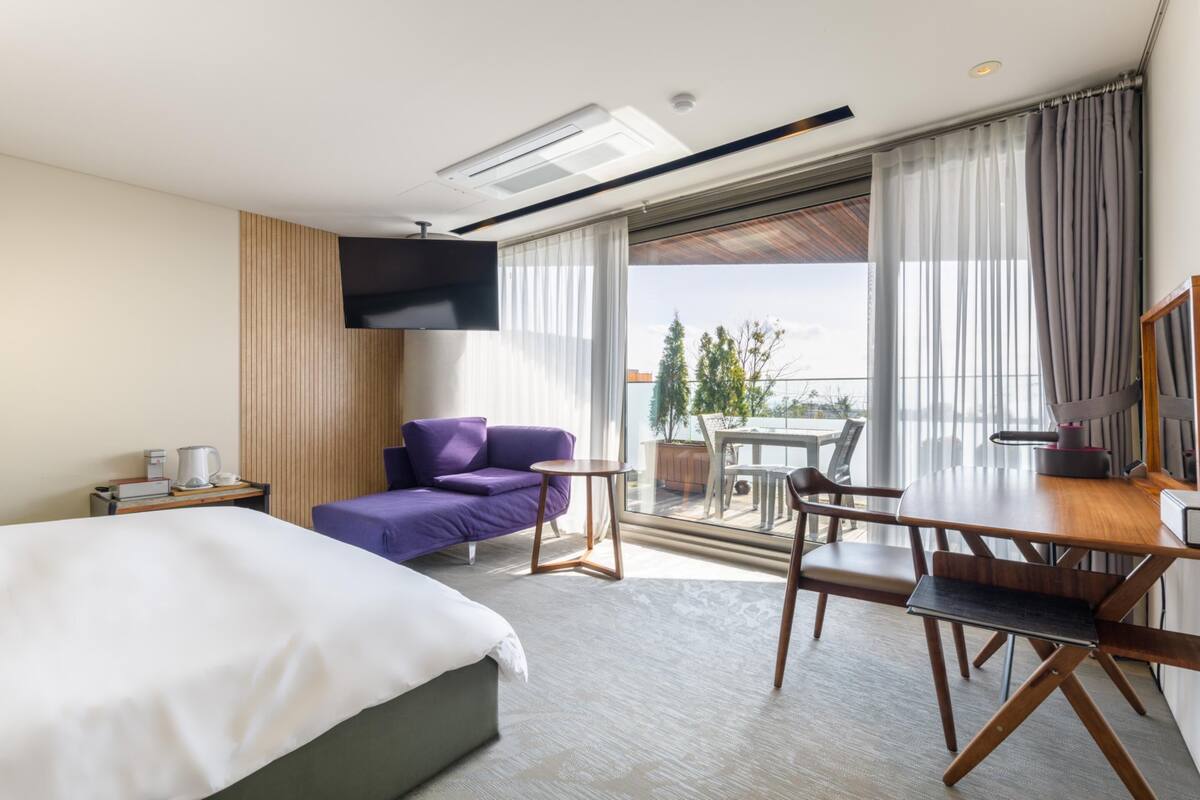 A modern hotel room with a large bed, a purple sofa, a dining table with chairs, a TV, and a balcony with a view of trees and buildings.