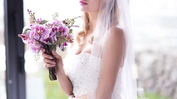 A bride in a white wedding dress holding a bouquet, seated at a table with a view of the outdoors.