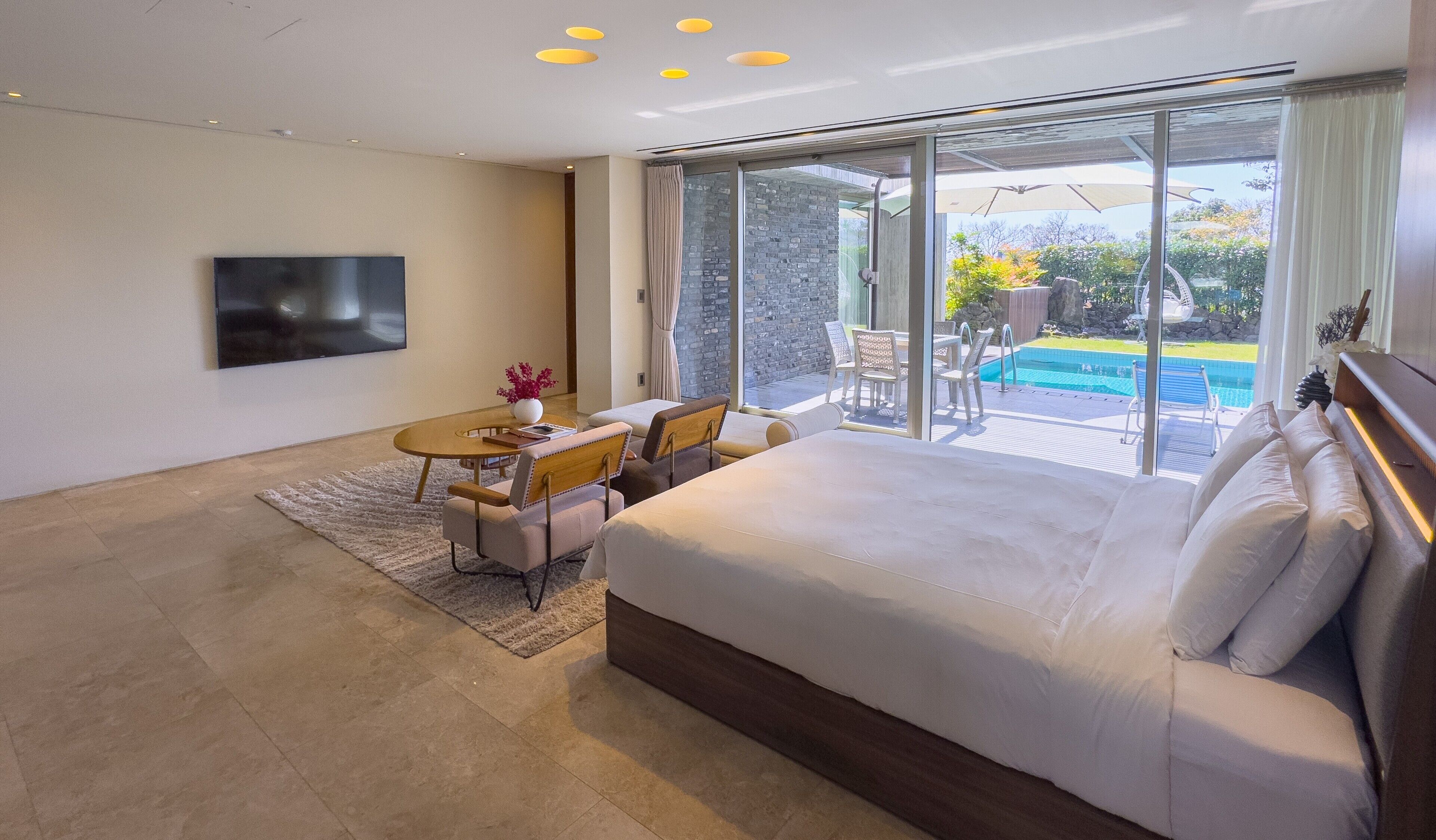 A modern hotel room with a large bed, a flat-screen TV, a dining area with a table and chairs, and a view of an outdoor swimming pool.
