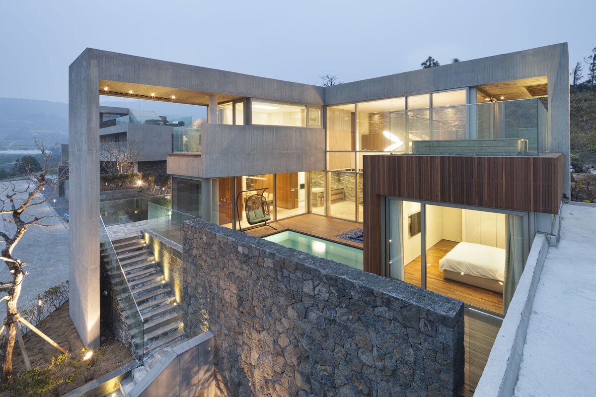 A modern house with a glass-enclosed swimming pool, a wooden deck, and a stone wall.