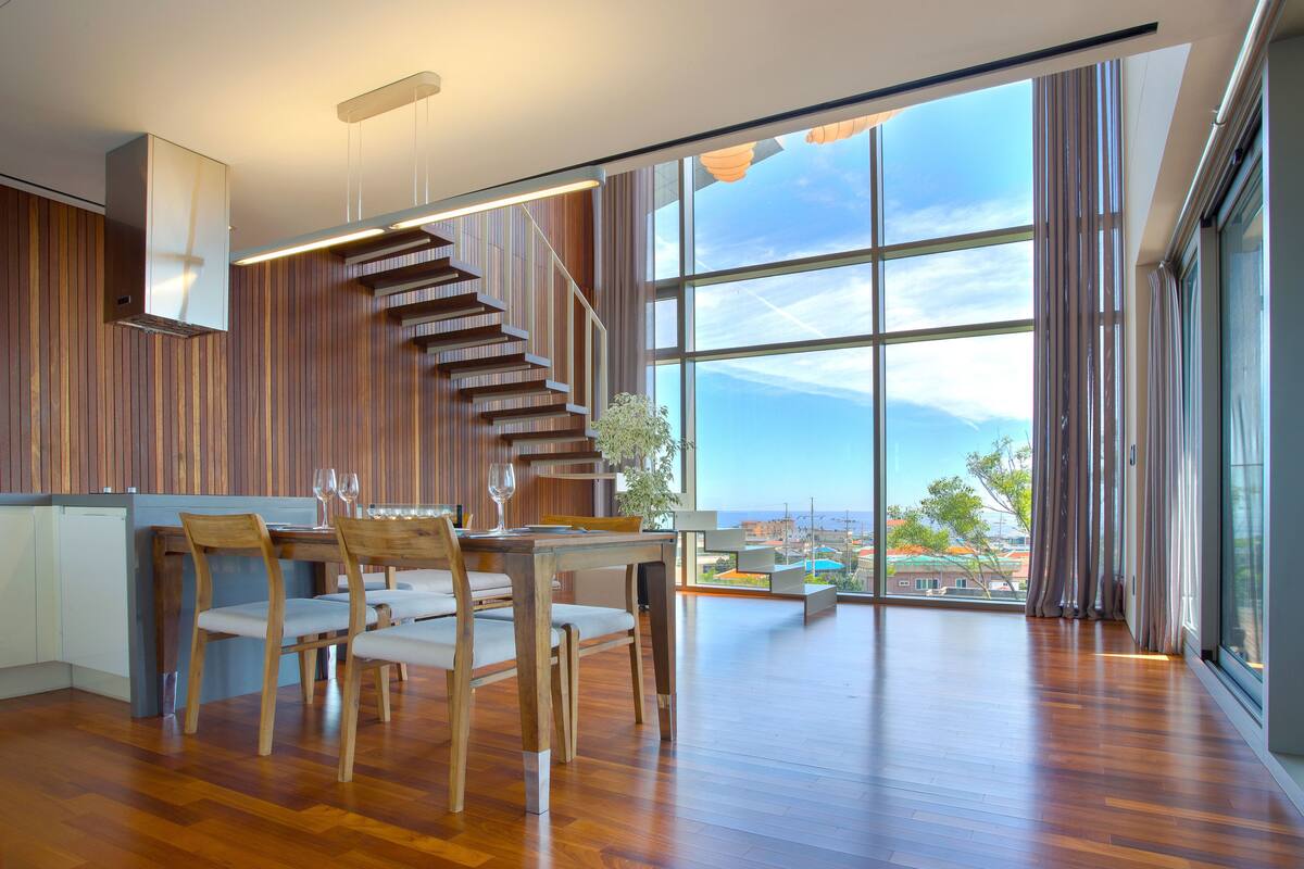 A modern interior with a wooden staircase, a dining area with chairs and a table, and large windows offering a view of the city.