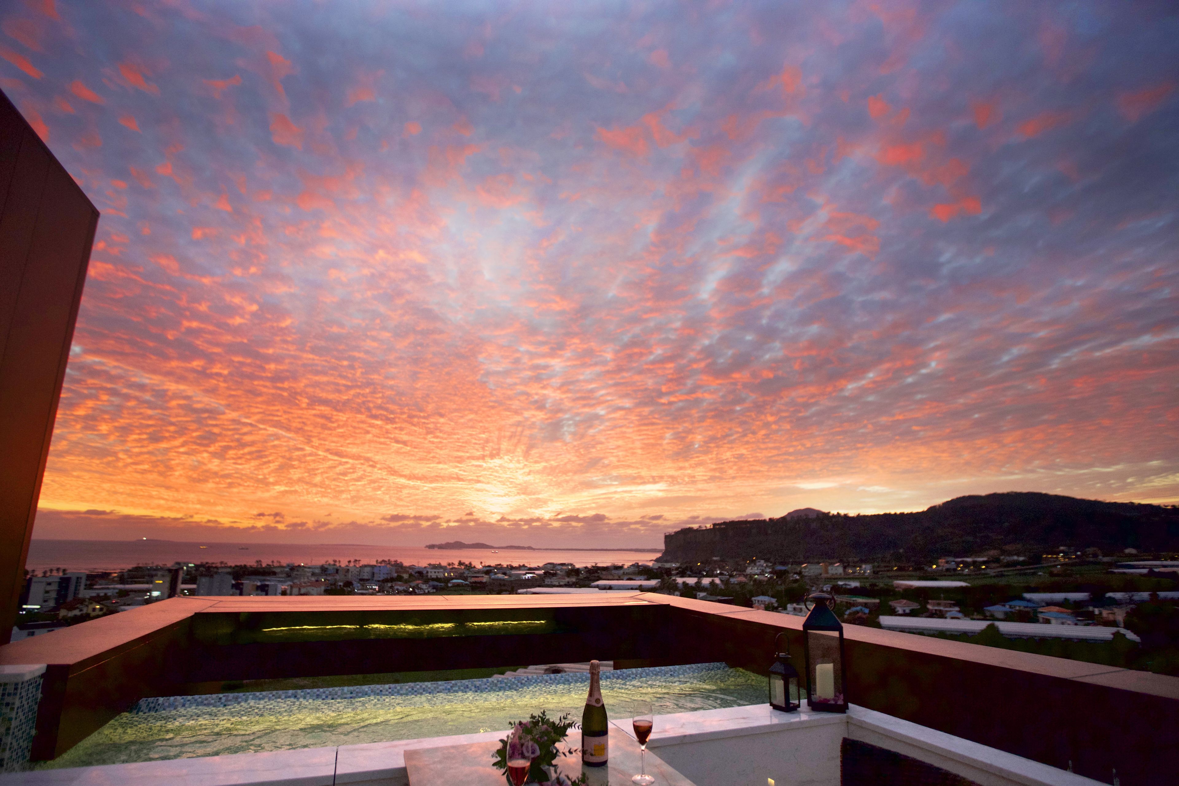 A rooftop terrace with a sunset view, a pool, and a table with a bottle and glass.
