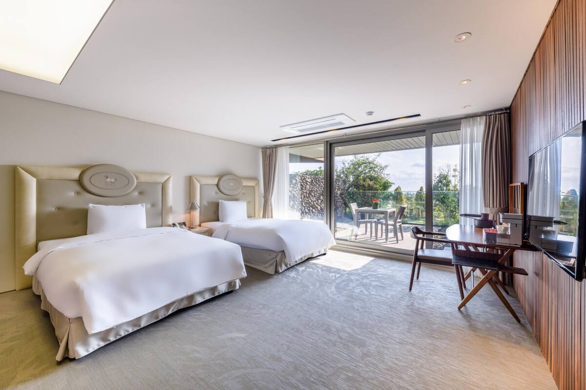 A modern hotel room with two beds, a dining area with a table and chairs, and a large window offering a view of the outdoors.