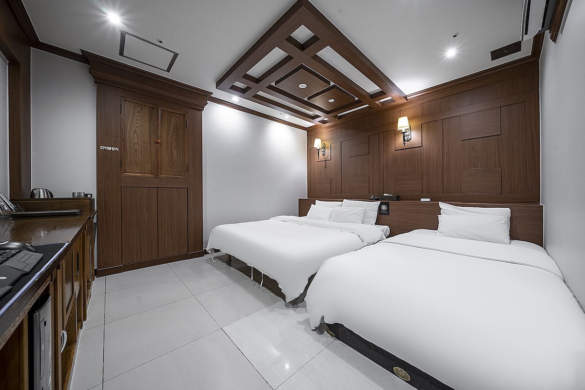 Une chambre d’hôtel avec deux lits, une armoire en bois, un bureau avec un ordinateur portable et un design moderne au niveau du plafond.