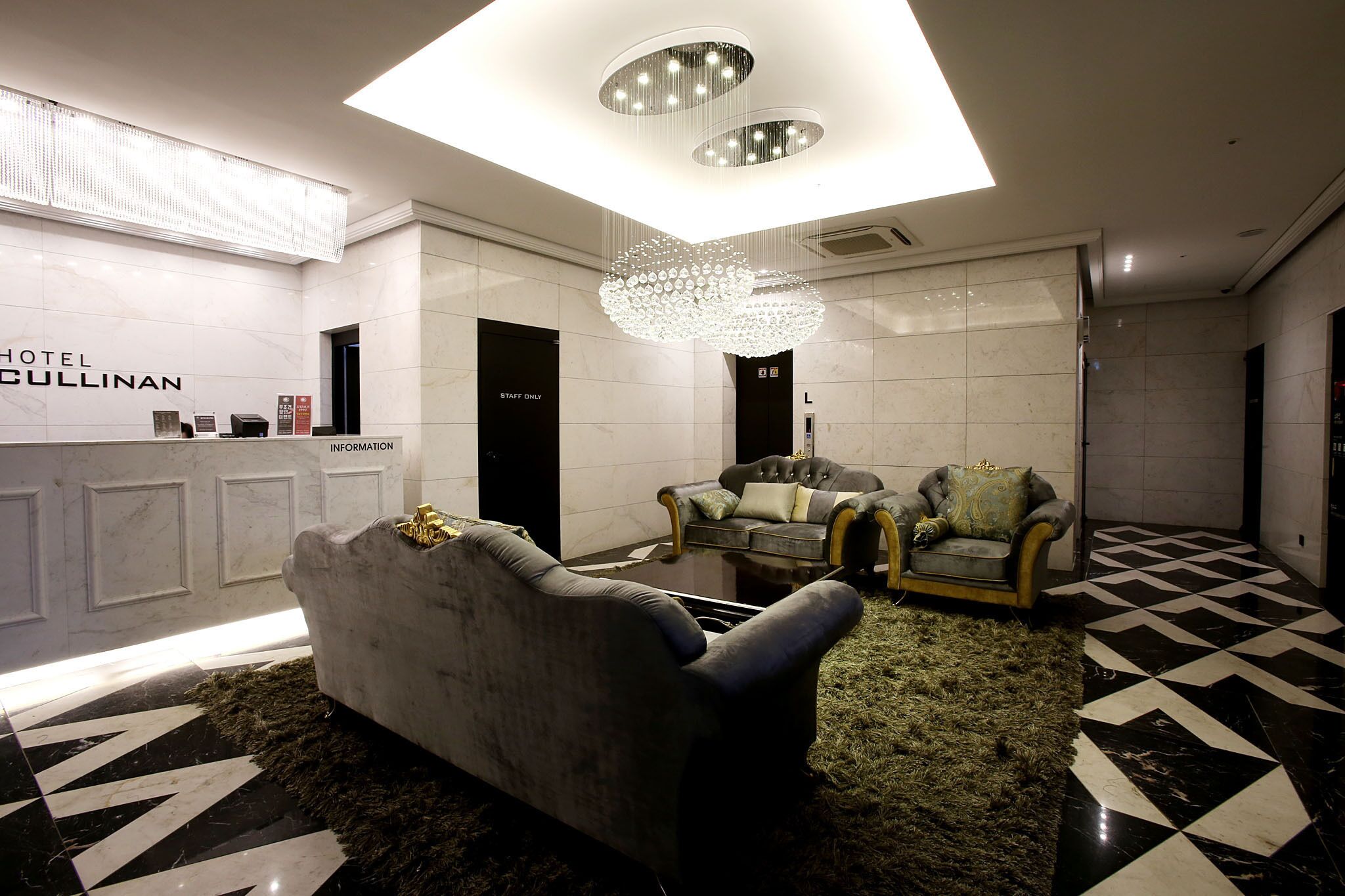 A hotel lobby with a reception desk, two armchairs, a chandelier, and a check-in counter.