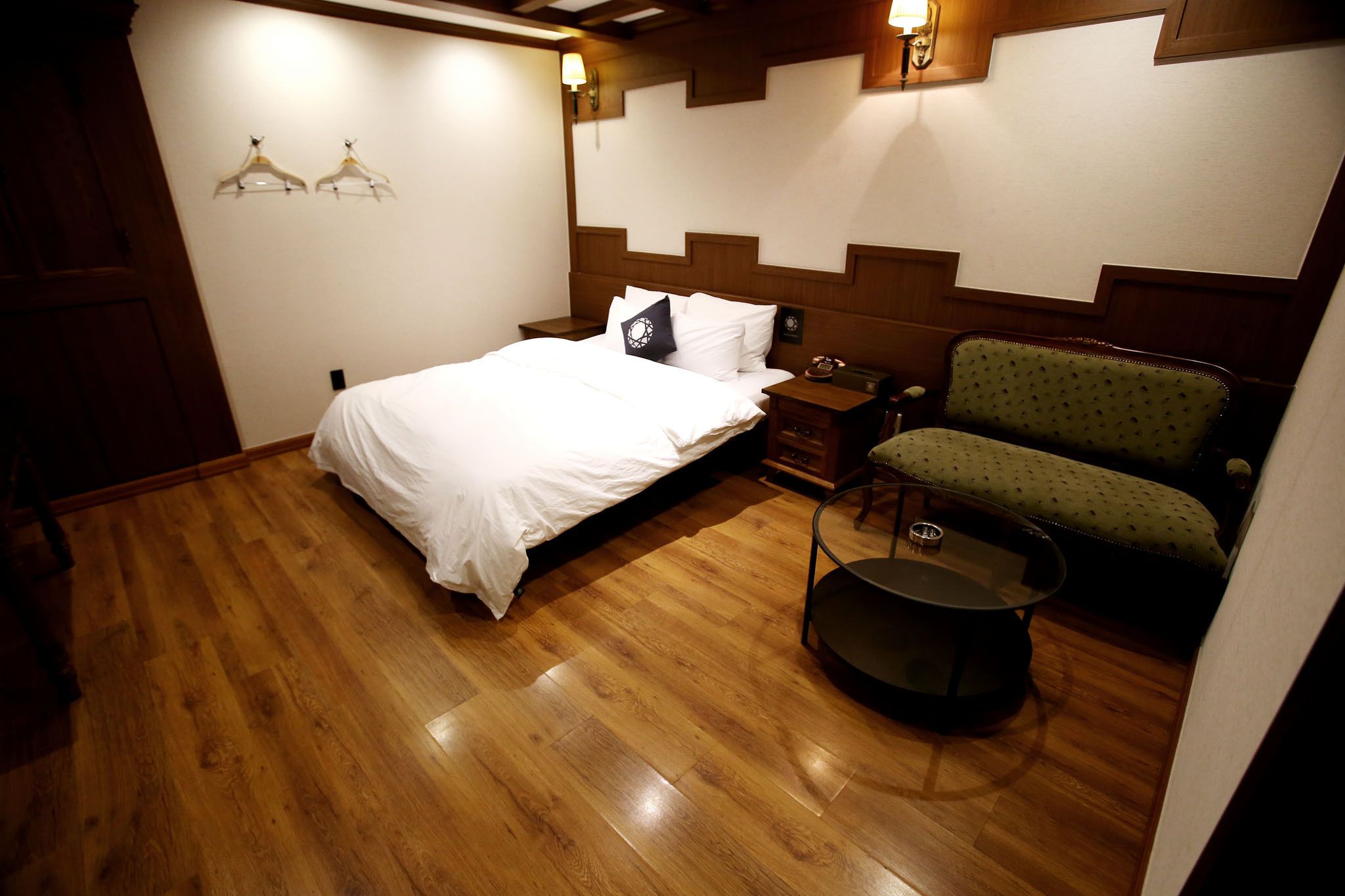A bedroom with a wooden floor, a bed with white bedding, a green upholstered chair, a small table, and wall-mounted lamps.