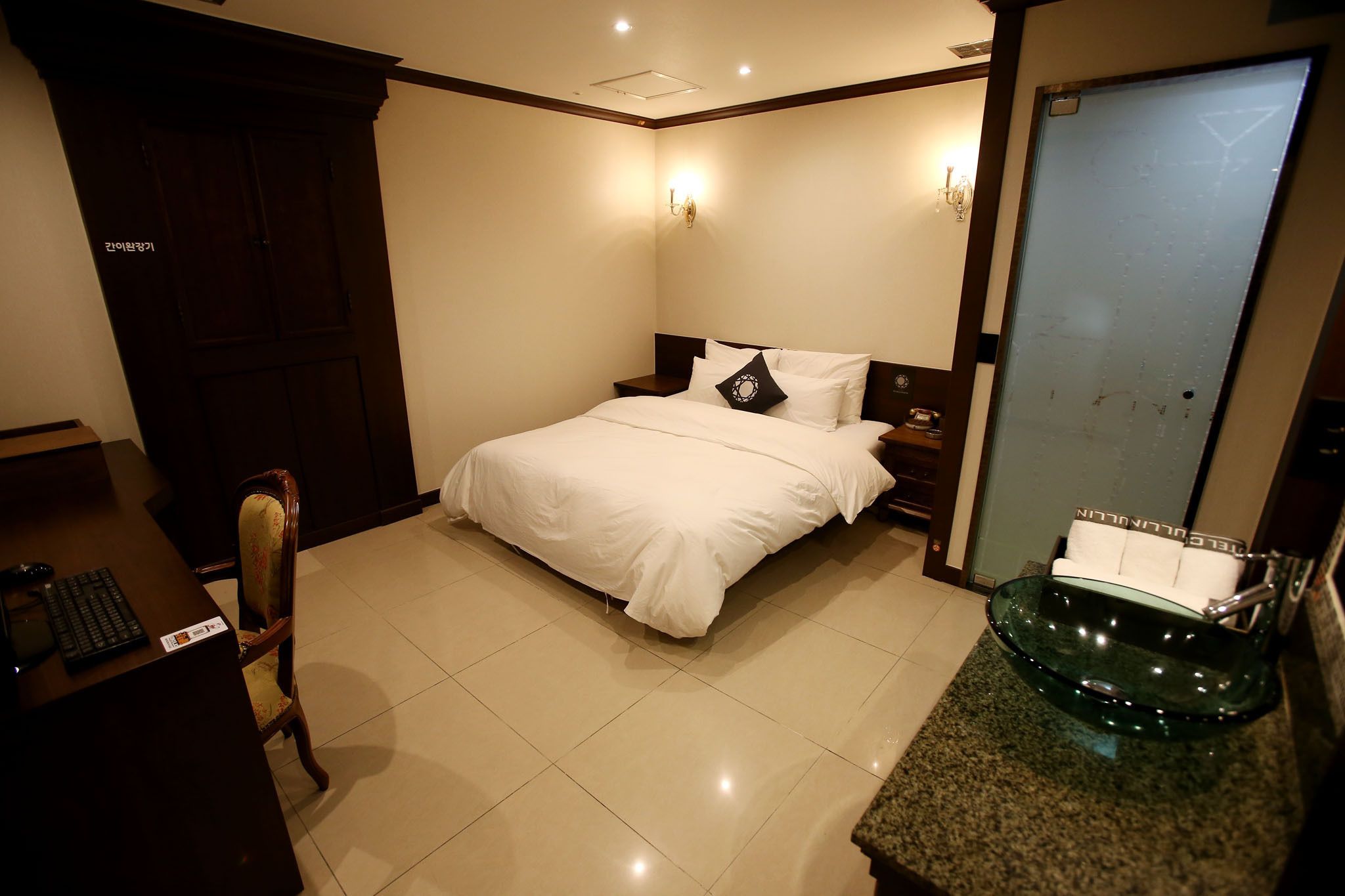 A hotel room with a bed, a desk with a keyboard, a chair, a glass sink, and a wardrobe.