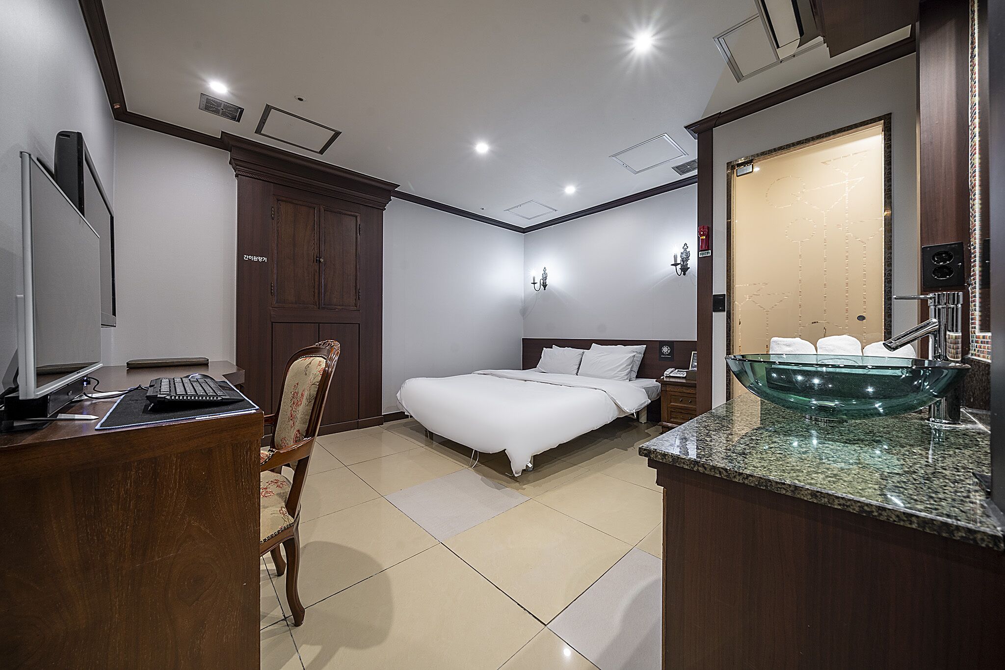 A hotel room with a bed, a desk with a computer, a chair, a nightstand, and a bathroom with a glass bowl sink.