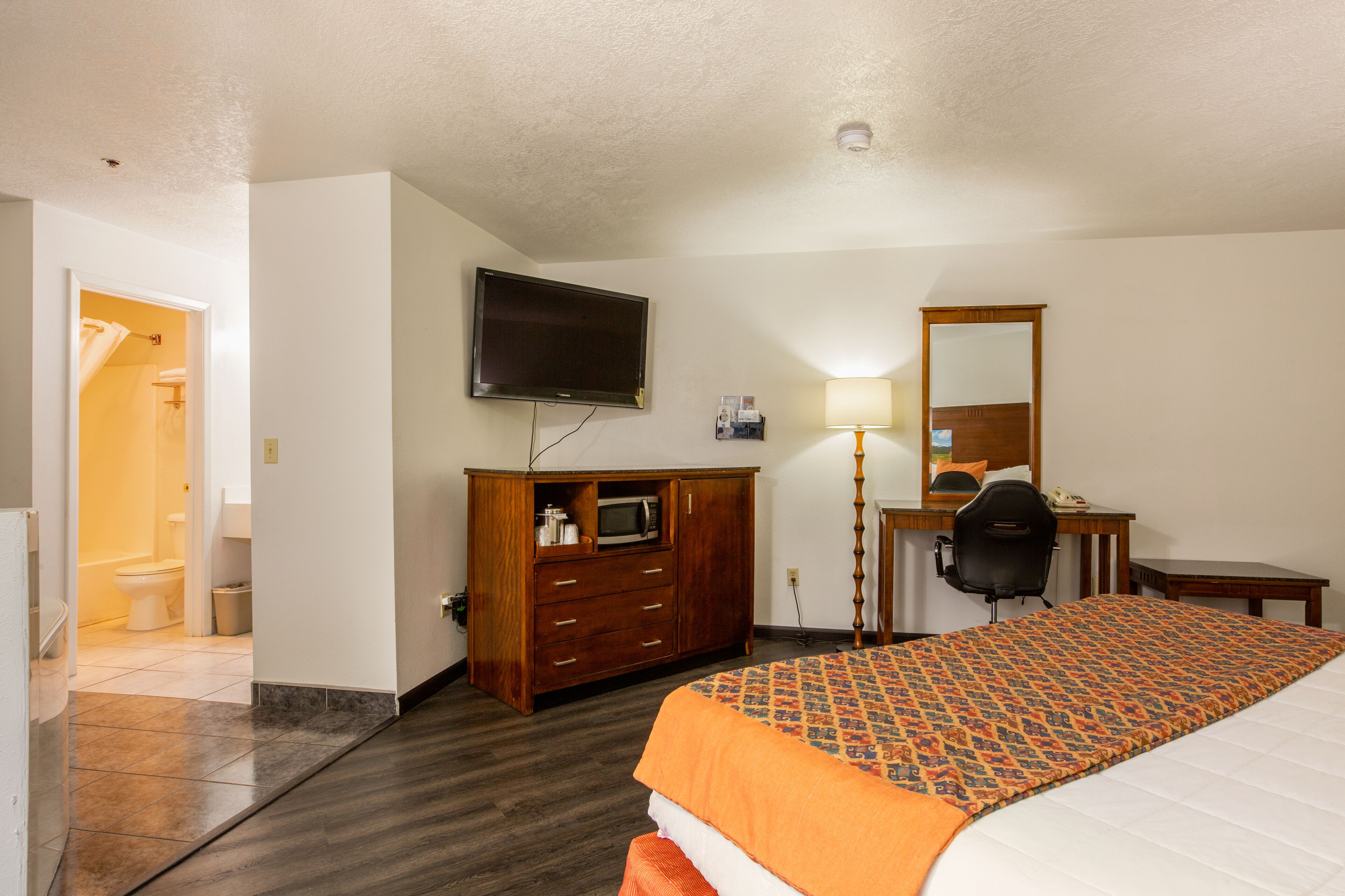 A hotel room with a bed, a desk with a chair, a television, a mirror, and a bathroom visible through an open door.