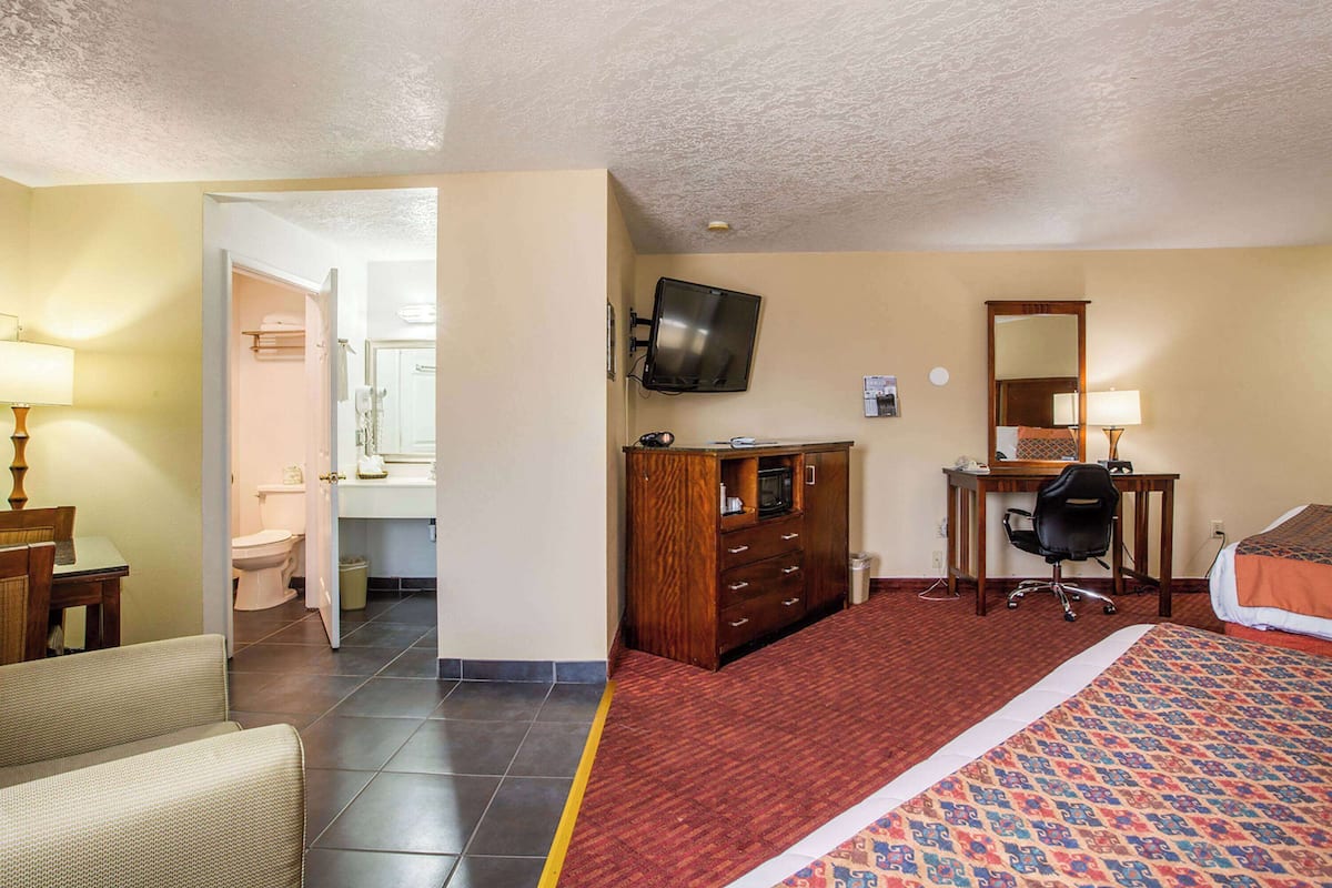 A hotel room with a bed, a desk with a chair, a television mounted on the wall, and a bathroom visible through an open door.
