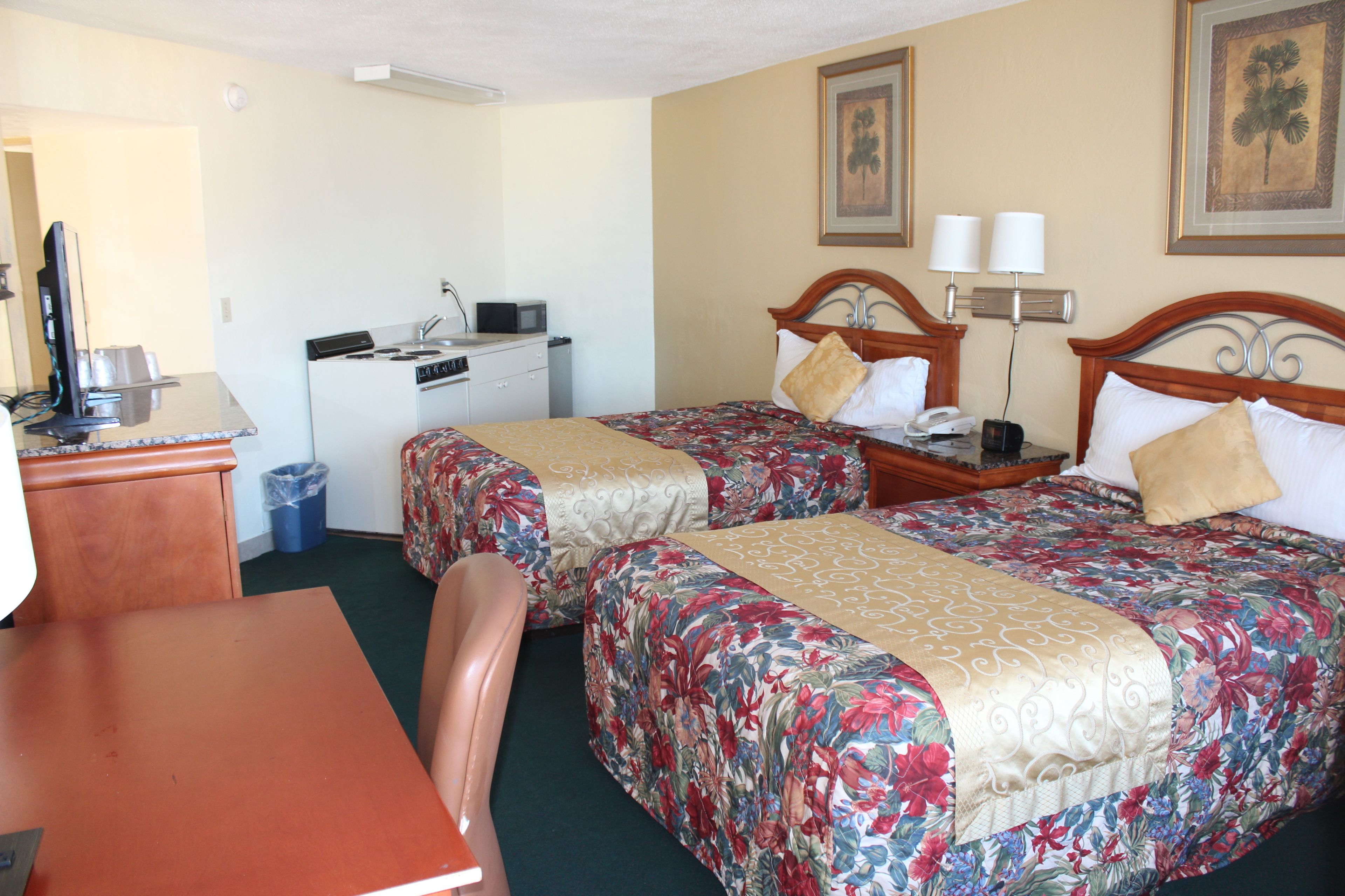 A hotel room with two beds, a desk, a chair, a small kitchenette, and a trash can.