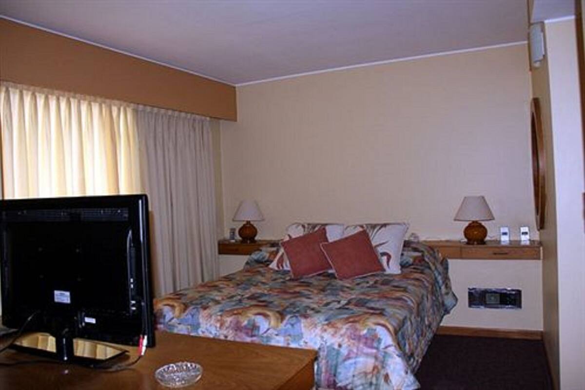 Quarto de hotel com cama, televisão, escrivaninha e abajur.