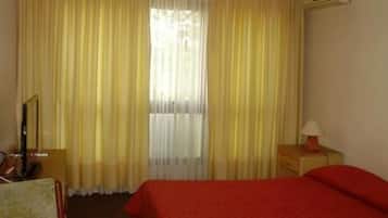 Habitación de hotel con cama, una mesita de noche con lámpara, una silla y un ventanal con cortinas.