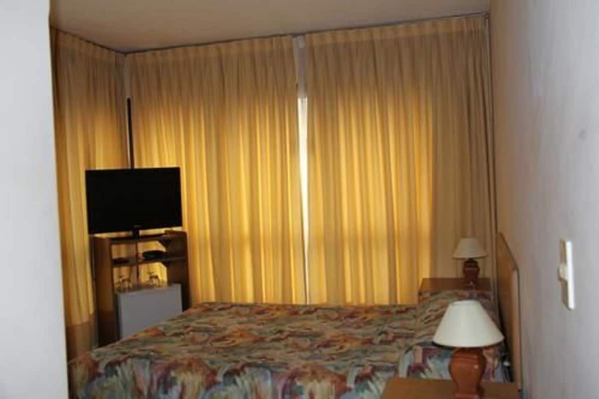 Quarto de hotel com uma cama, televisão em suporte, luminária e cortinas.