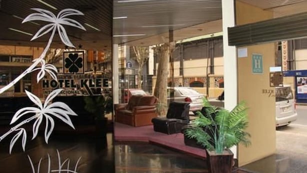 Entrada de hotel com balcão de recepção, área de estar com cadeiras e um vaso de planta.