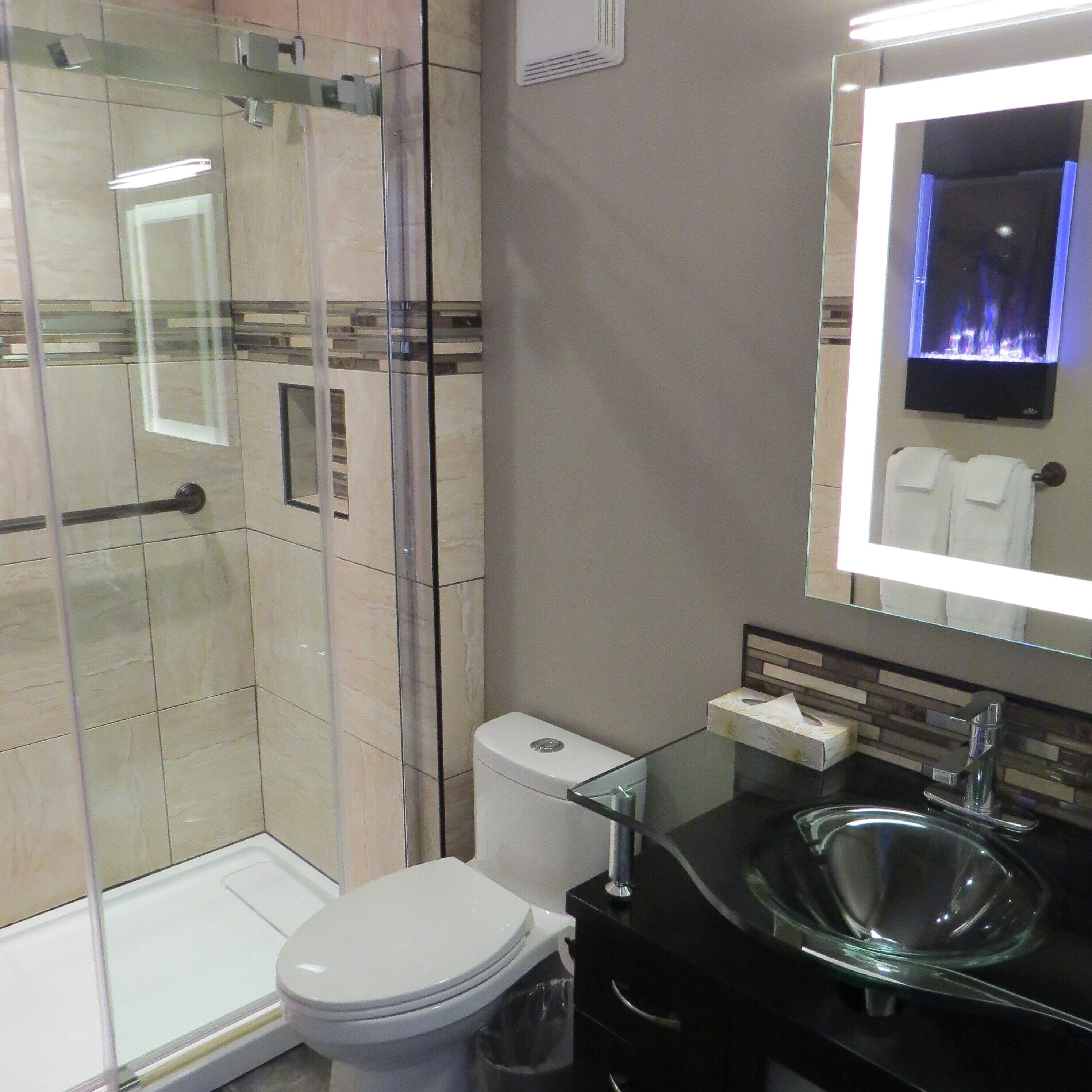 Une salle de bain moderne avec une douche à l’italienne en verre, des toilettes blanches, un lavabo noir avec un receveur en verre et un miroir doté d’un éclairage arrière bleu.