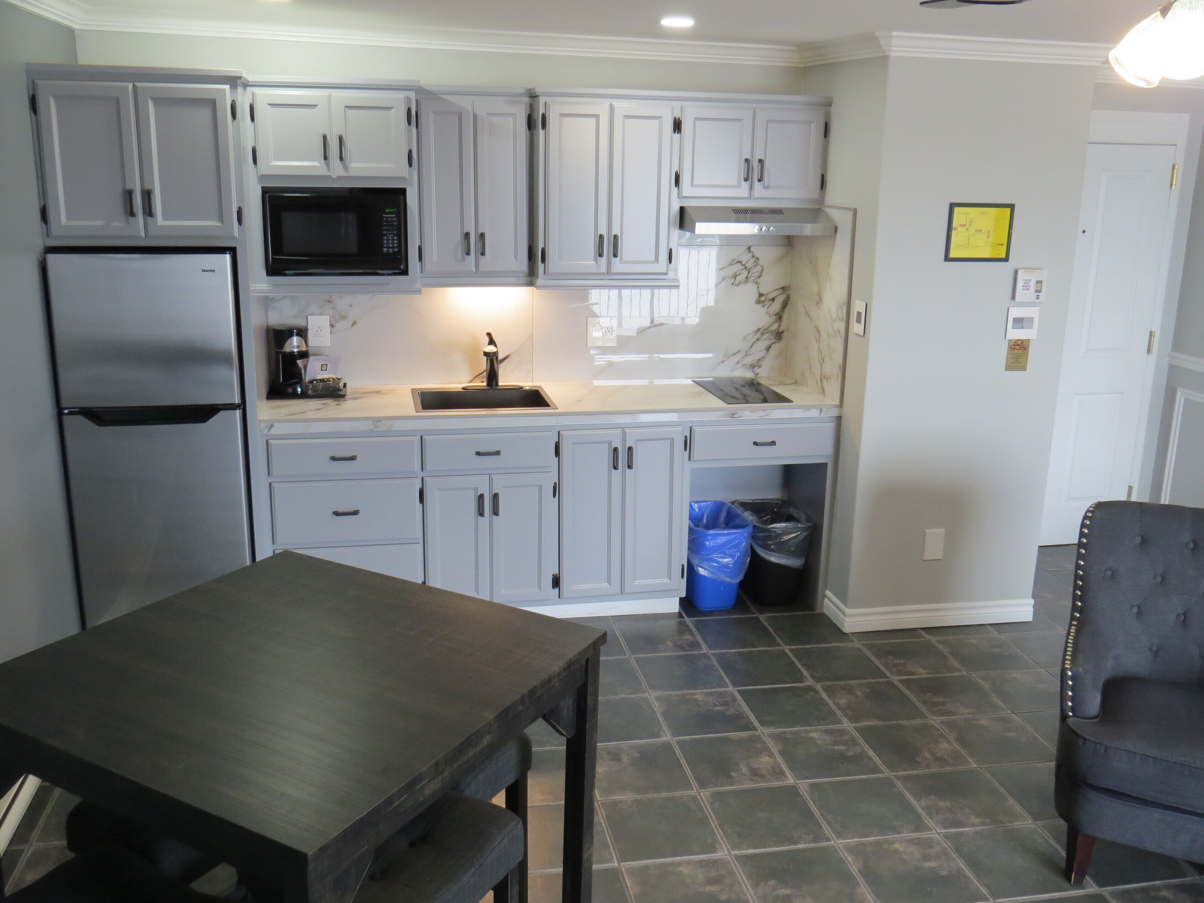 A kitchen with grey cabinets, a stainless steel refrigerator, a microwave, and a sink. There is a blue trash bin under the counter.