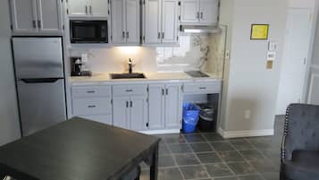A kitchen with grey cabinets, a stainless steel refrigerator, a microwave, and a sink. There is a blue trash bin under the counter.